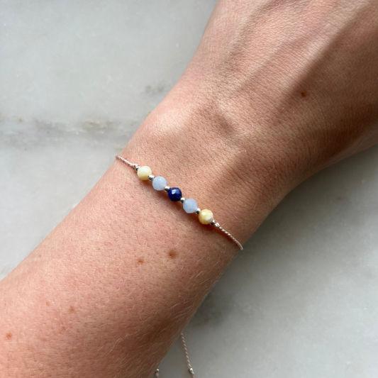 A hand modeling pink silk cord bracelet strung with these mineral beads: yellow opal, blue lapis lazuli, blue aquamarine. Between each mineral is also a little silver bead separating the minerals and making the design more elegant.