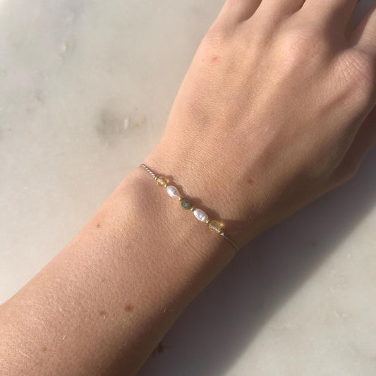 A female hand modeling a grey silk cord bracelet strung with these mineral beads: yellow citrine, white pearl, green emerald, another pearl and another citrine. Between the minerals, there are also little gold beads separating them and making the design more elegant.