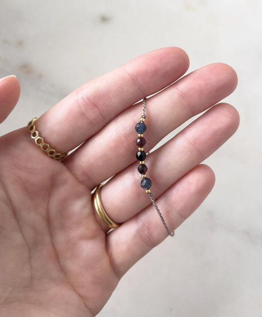 A delicate beaded bracelet with small, colourful gemstones. The bracelet features dark blue, burgundy, and gold-coloured beads. The bracelet is held in a hand, with gold rings on the fingers.