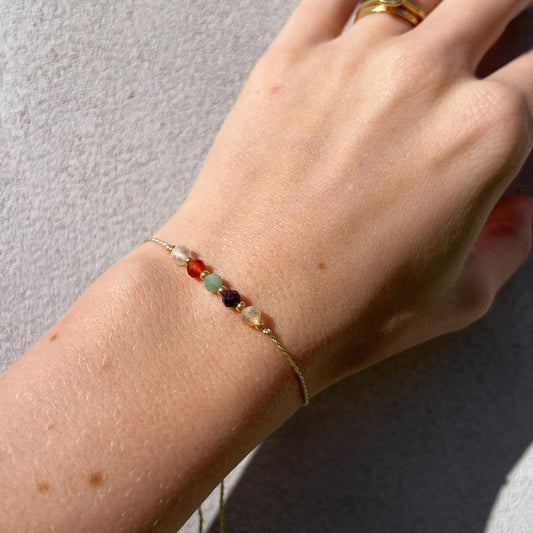 A female hand modeling a green silk cord bracelet strung with five mineral beads: yellow citrine, red garnet, red carnelian, clear quartz, green emerald. Each mineral is separated from another with a little gold bead, making the design more elegant. Model is also wearing a dainty gold bracelet together with the mineral bracelet, showing how the two can be elegantly layered together.