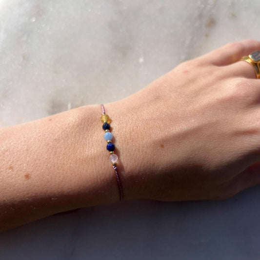 A hand modeling lilac silk cord bracelet strung with these mineral beads: yellow opal, blue lapis lazuli, blue aquamarine, another lapis lazuli, rose quartz. Between each mineral is also a little gold bead separating the minerals and making the design more elegant.