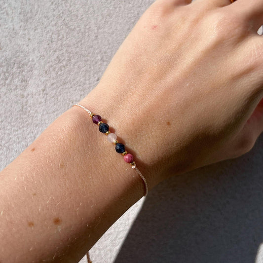 A female hand modeling a pink silk cord bracelet strung with five mineral beads: pink rhodonite, blue sapphire, rose quartz, blue sapphire, purple amethyst. Each mineral is separated from another with a little gold bead, making the design more elegant.