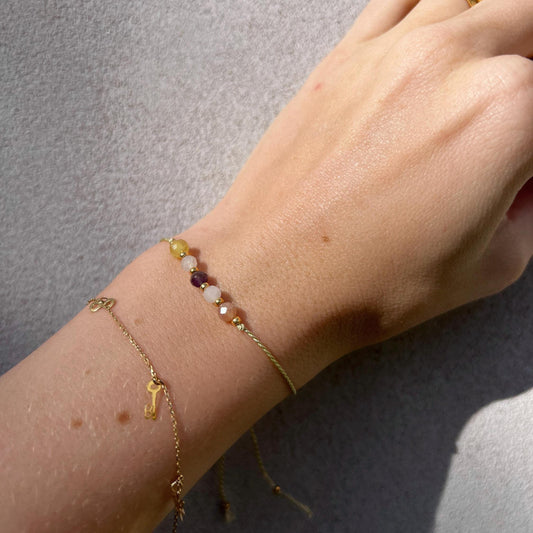 A female hand modeling a green silk cord bracelet strung with five mineral beads: purple amethyst, white moonstone, yellow opal, orange sunstone, rose quartz. Each mineral is separated from another with a little gold bead, making the design more elegant. Model is also wearing a dainty gold bracelet together with the mineral bracelet, showing how the two can be elegantly layered together.