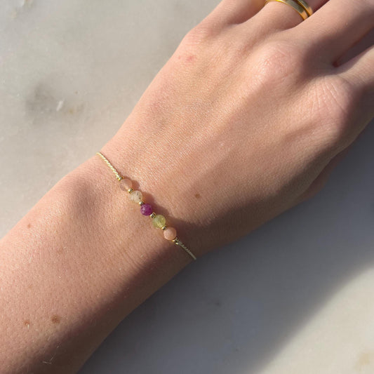 A female hand modeling a green silk cord bracelet strung with these mineral beads: orange sunstone, green peridot, red ruby, yellow citrine and another sunstone. Between the minerals, there are also little gold beads separating them and making the design more elegant.