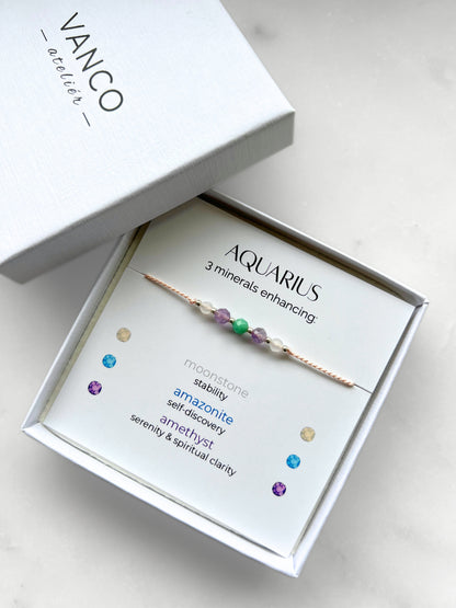 White gift box that contains a mineral bracelet for an Aquarius Zodiac sign. The bracelet is displayed on a card that describes the minerals and their meanings. There are five minerals on the bracelet: two moonstones, two amazonites and amethyst