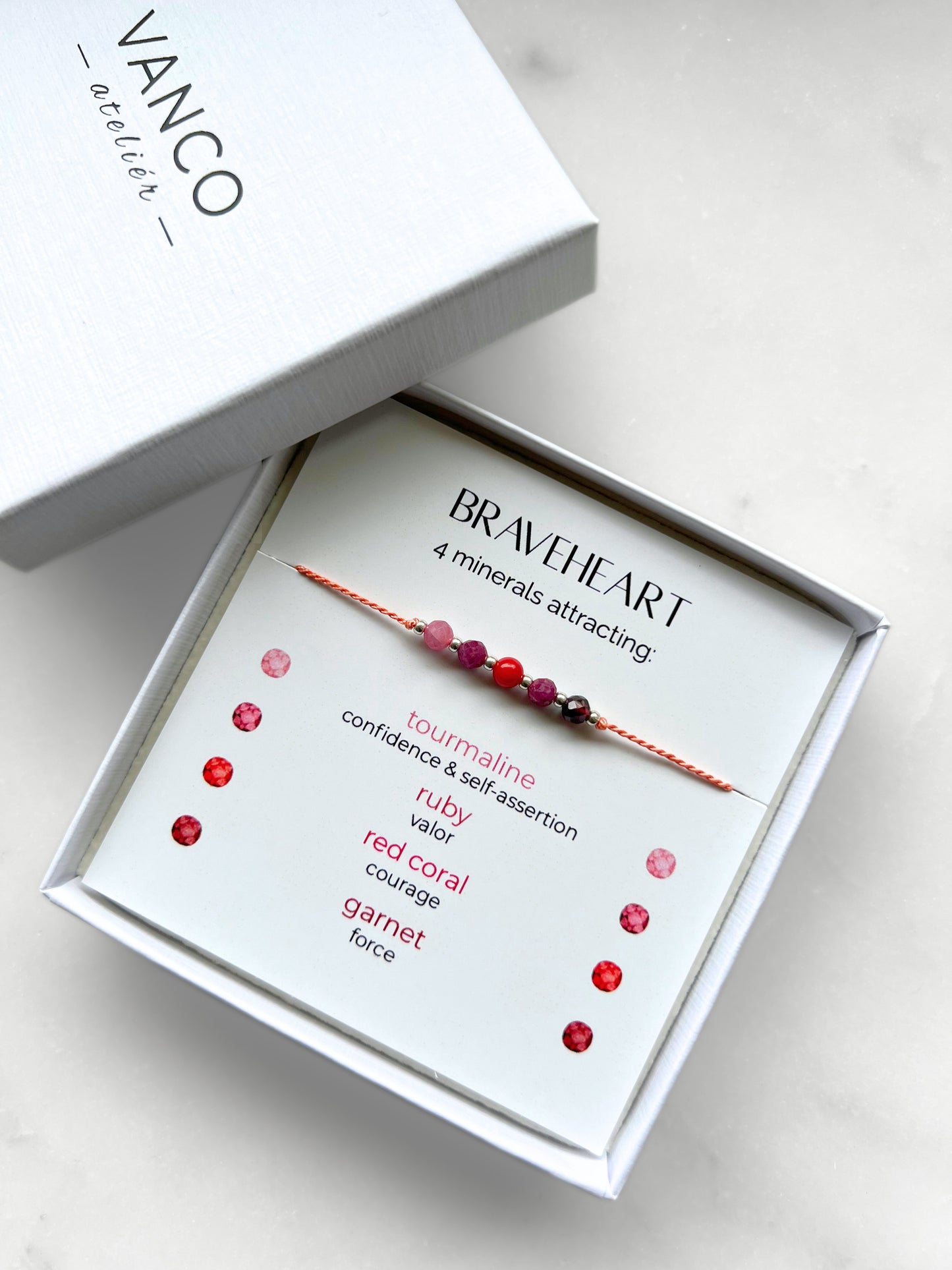 A white gift box that contains a bracelet made of tangerine silk string with five mineral beads, put on a promotional card. The bracelet's theme is Courage, the name of the bracelet is Braveheart which is written in the promotional card. The card also explains each stone and what each stone represents: Red Coral for Courage, Ruby for Valor, Garnet for Force, Pink Tourmaline for Confidence & Self-assertion