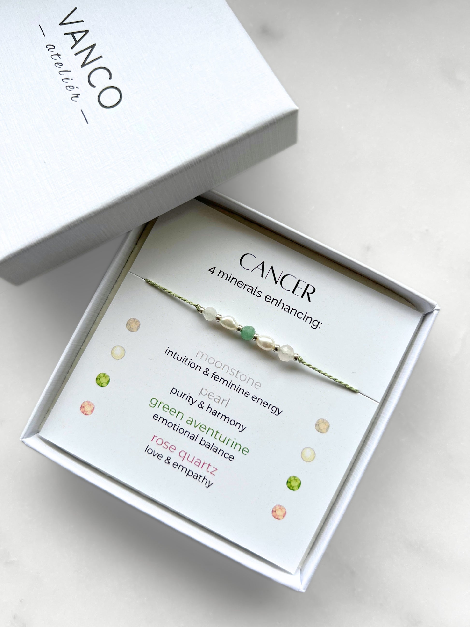 White gift box that contains a mineral bracelet for a Cancer Zodiac sign. The bracelet is displayed on a card that describes the minerals and their meanings. There are five minerals on the bracelet: moonstone, two pearls, green aventurine and rose quartz
