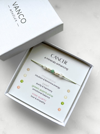 White gift box that contains a mineral bracelet for a Cancer Zodiac sign. The bracelet is displayed on a card that describes the minerals and their meanings. There are five minerals on the bracelet: moonstone, two pearls, green aventurine and rose quartz