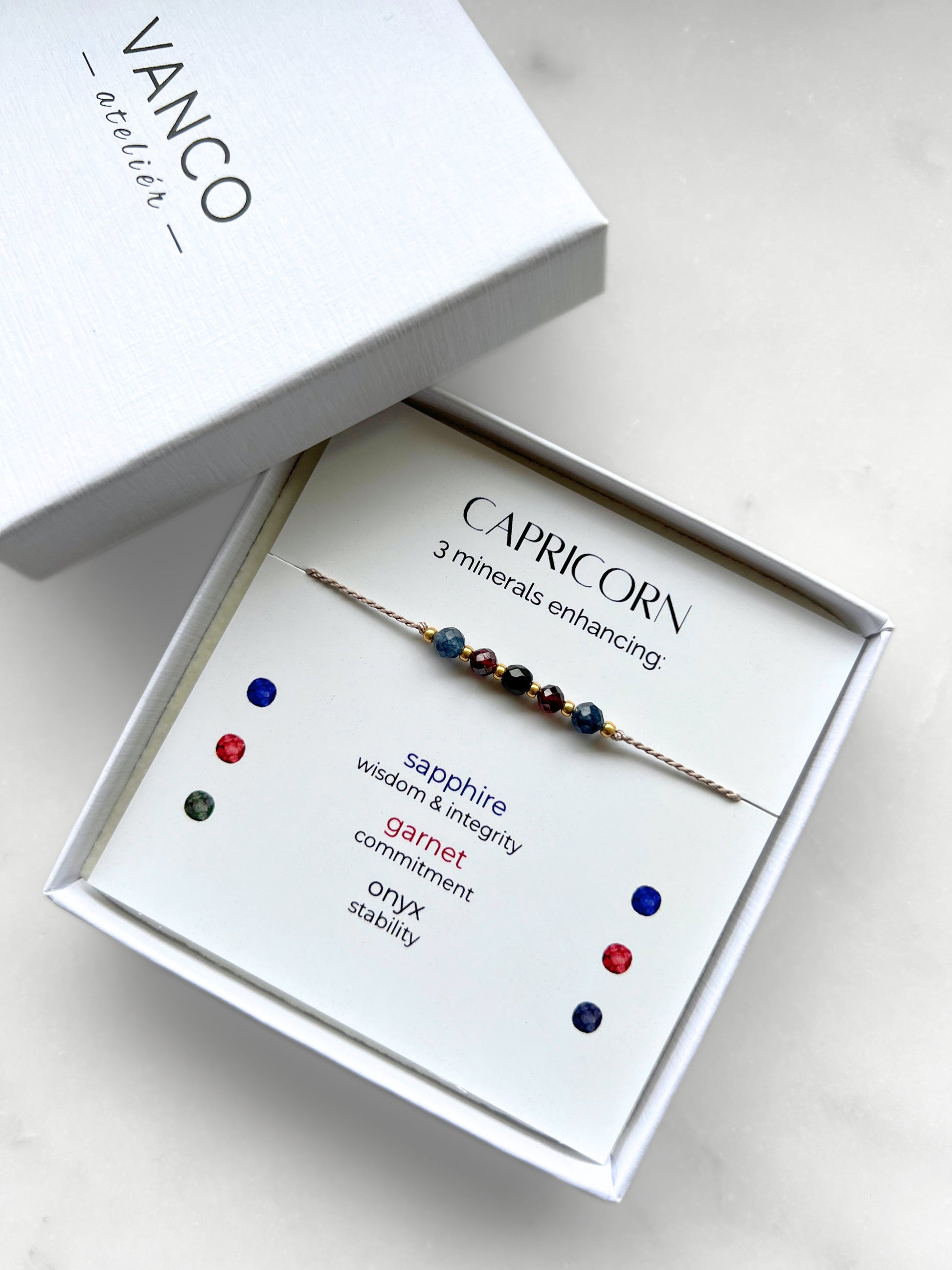 White gift box that contains a mineral bracelet for a Capricorn Zodiac sign. The bracelet is displayed on a card that describes the minerals and their meanings. There are five minerals on the bracelet: two sapphires, two garnets and onyx