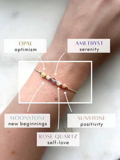 A female hand modeling a green silk cord bracelet strung with five mineral beads: purple amethyst, white moonstone, yellow opal, orange sunstone, rose quartz. Each mineral is separated from another with a little gold bead, making the design more elegant. Model is also wearing a dainty gold bracelet together with the mineral bracelet, showing how the two can be elegantly layered together.