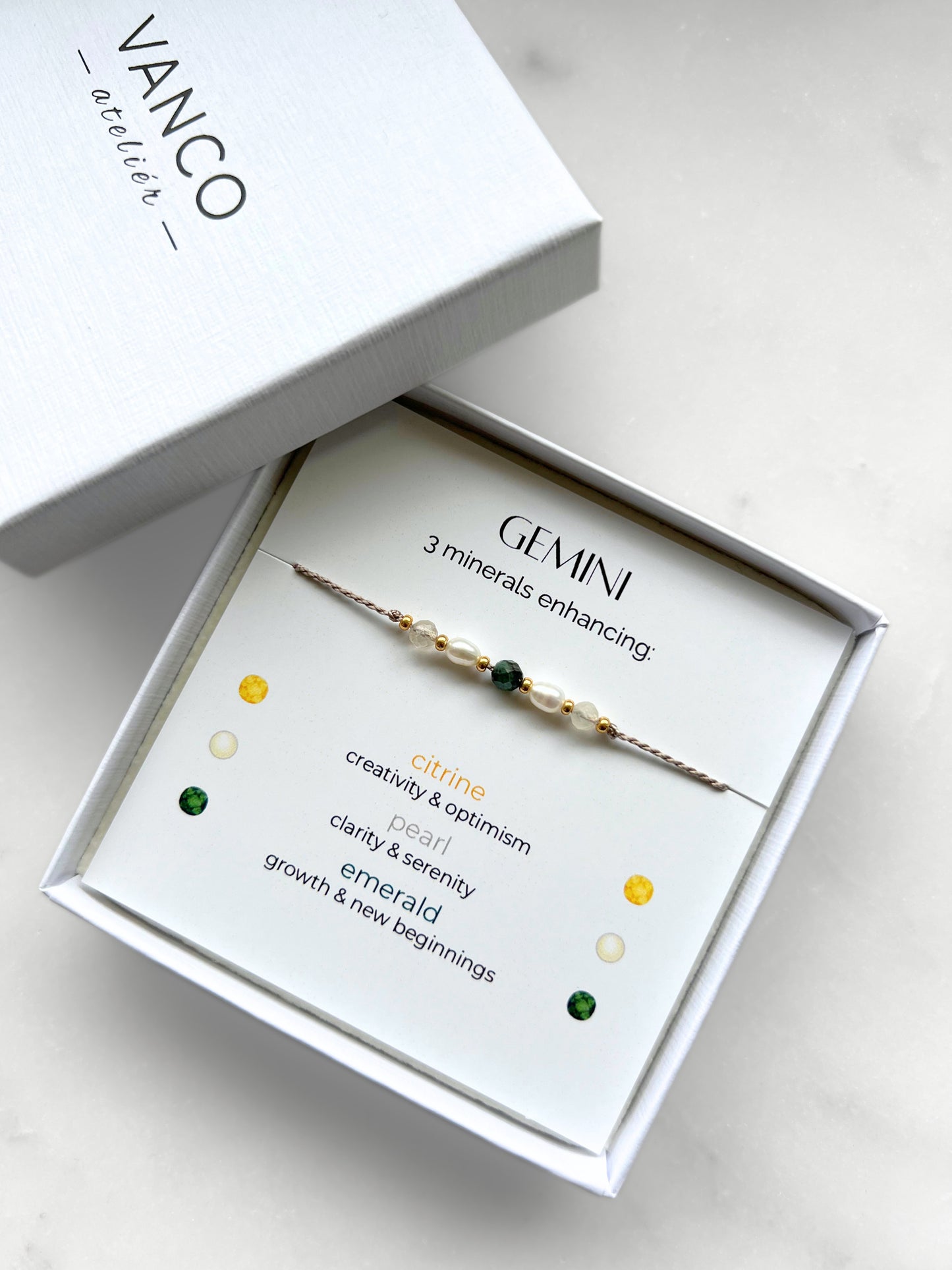 White gift box that contains a mineral bracelet for a Gemini Zodiac sign. The bracelet is displayed on a card that describes the minerals and their meanings. There are five minerals on the bracelet: two citrines, two pearls and one emerald