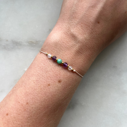 A female hand modeling a lilac silk cord bracelet strung with these mineral beads: white moonstone, blue amazonite, purple amethyst. Between the minerals, there are also little gold beads separating them and making the design more elegant.