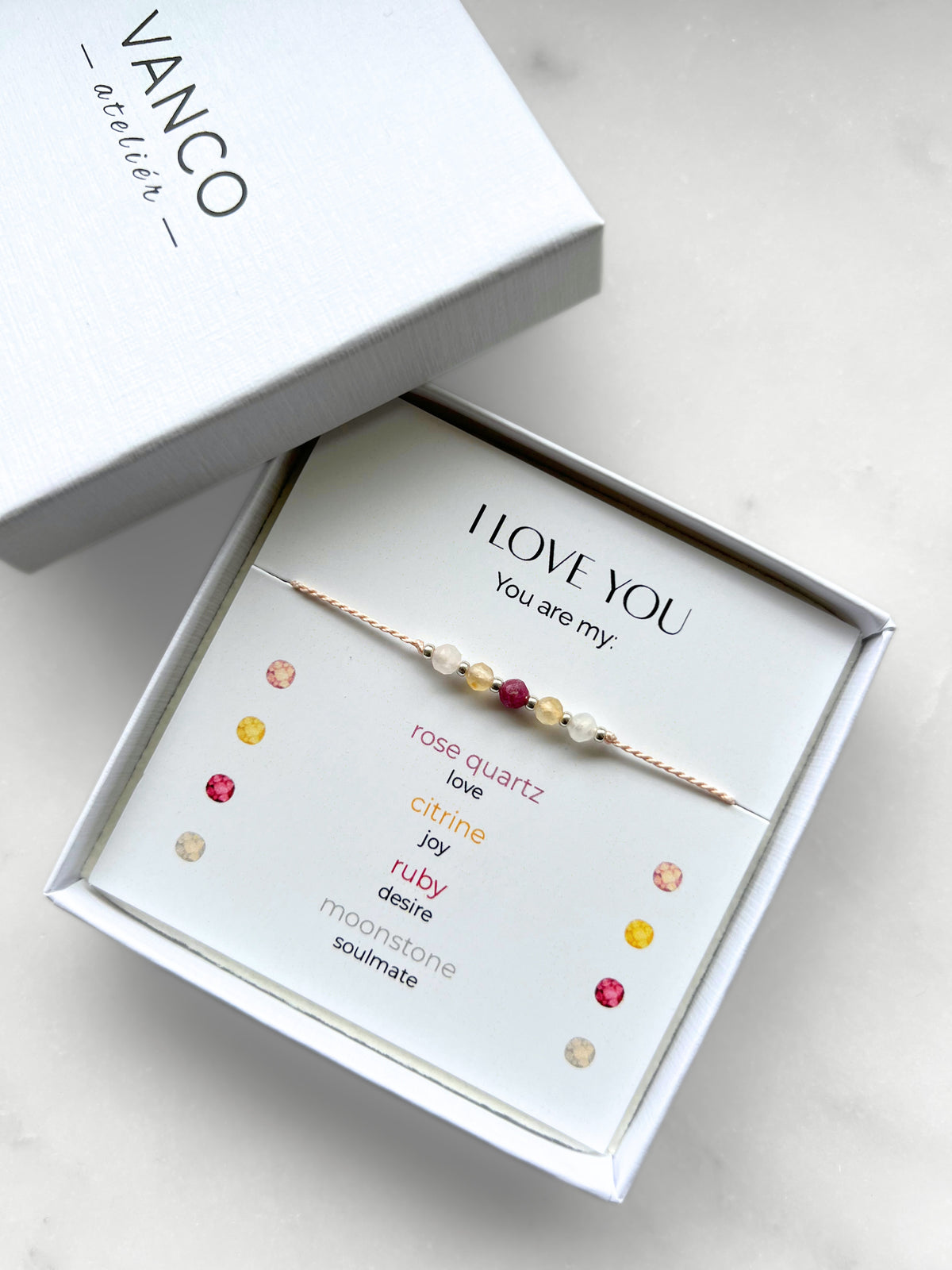 A white gift box that contains a bracelet made of pink silk string with five mineral beads, put on a promotional card. The bracelet's theme is "Love Declaration", the name of the bracelet is "I Love You", which is written in the promotional card. The card also explains each stone and what message each stone sends to the recipient, saying "Your are my": Rose Quartz for Love, Citrine for Joy, Ruby for Desire and Moonstone for Soulmate.
