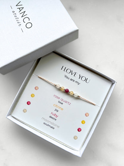 A white gift box that contains a bracelet made of pink silk string with five mineral beads, put on a promotional card. The bracelet's theme is "Love Declaration", the name of the bracelet is "I Love You", which is written in the promotional card. The card also explains each stone and what message each stone sends to the recipient, saying "Your are my": Rose Quartz for Love, Citrine for Joy, Ruby for Desire and Moonstone for Soulmate.