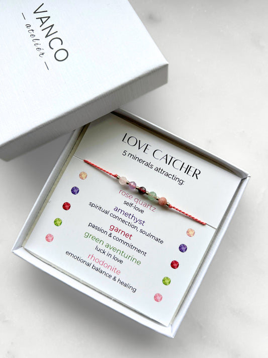 A white gift box that contains a bracelet made of pink silk string with five mineral beads, put on a promotional card. The bracelet's theme is Love, the name of the bracelet is Love Catcher which is written in the promotional card. The card also explains each stone and what each stone represents: Rose Quartz for Self-love, Amethyst for Spiritual Connection & Soulmate, Garnet for Passion & Commitment, Green Aventurine for Luck in Love, Rhodonite for Emotional Balance & Healing