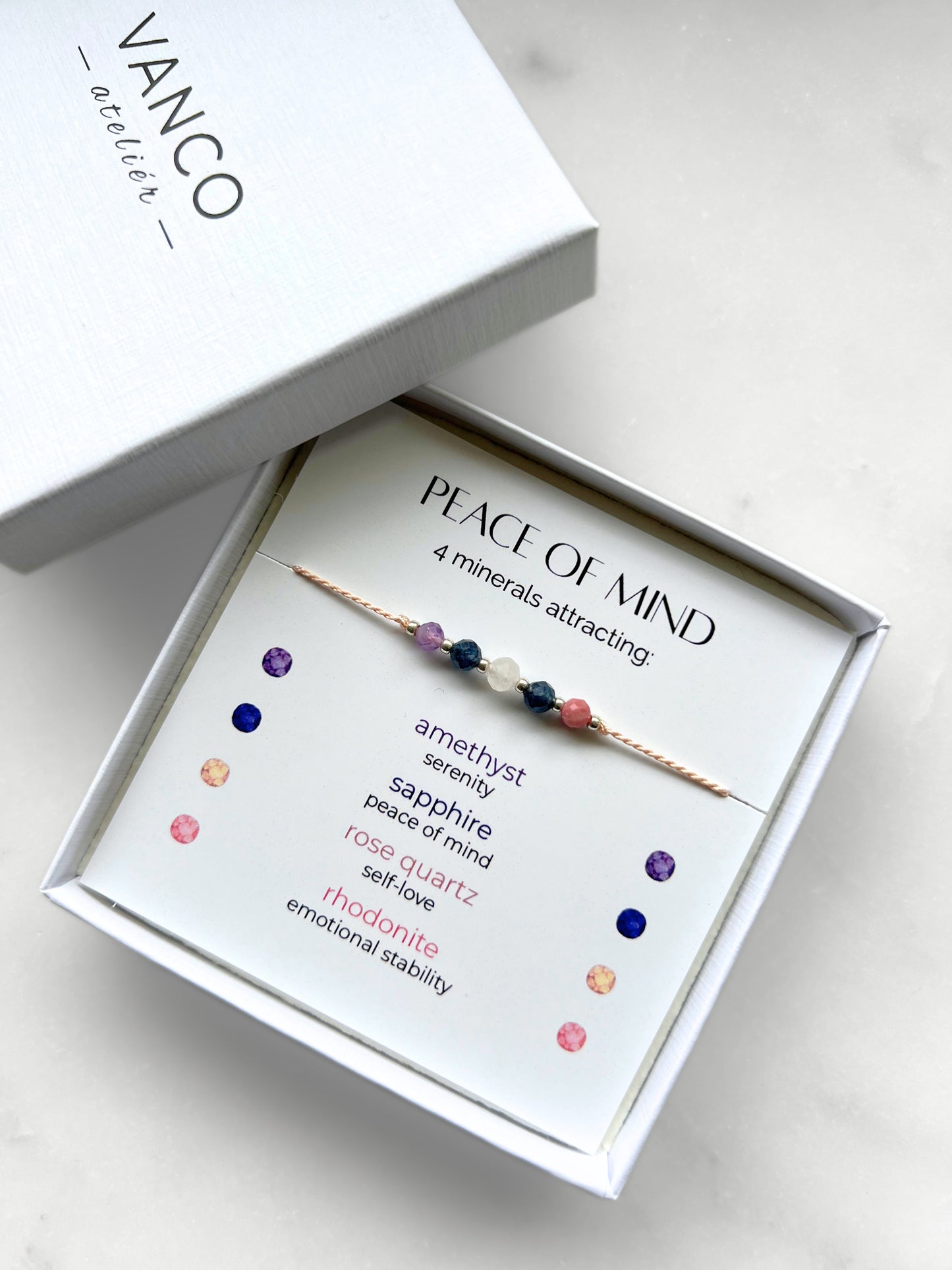 A white gift box that contains a bracelet made of pink silk string with five mineral beads, put on a promotional card. The bracelet's theme is Serenity, the name of the bracelet is Peace of Mind which is written in the promotional card. The card also explains each stone and what each stone represents: Amethyst for Serenity, Sapphire for Peace of Mind, Rose Quartz for Self-love, Rhodonite for Emotional Stability