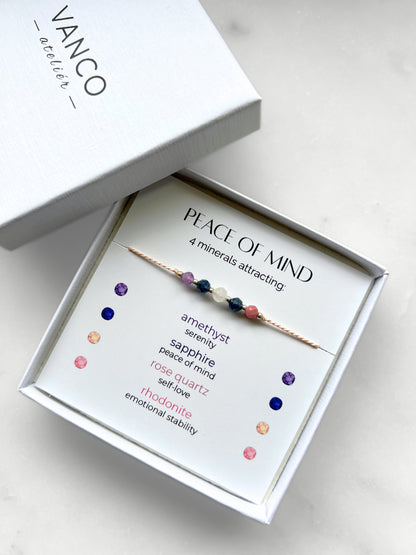 A white gift box that contains a bracelet made of pink silk string with five mineral beads, put on a promotional card. The bracelet's theme is Serenity, the name of the bracelet is Peace of Mind which is written in the promotional card. The card also explains each stone and what each stone represents: Amethyst for Serenity, Sapphire for Peace of Mind, Rose Quartz for Self-love, Rhodonite for Emotional Stability