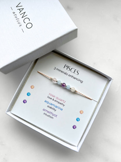 White gift box that contains a mineral bracelet for a Pisces Zodiac sign. The bracelet is displayed on a card that describes the minerals and their meanings. There are five minerals on the bracelet: two rose quartz, two aquamarines and amethyst