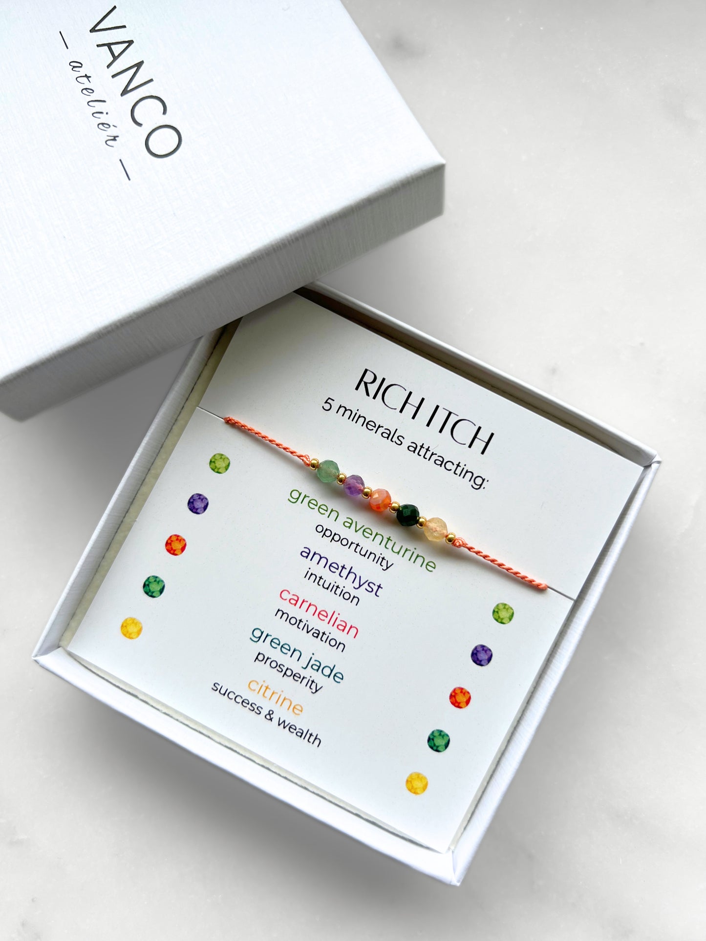 A white gift box that contains a bracelet made of tangerine silk string with five mineral beads, put on a promotional card. The bracelet's theme is Wealth, the name of the bracelet is Rich Itch which is written in the promotional card. The card also explains each stone and what each stone represents: Green Aventurine for Opportunity, Amethyst for Intuition, Carnelian for Motivation, Green Jade for Prosperity, Citrine for Success & Wealth