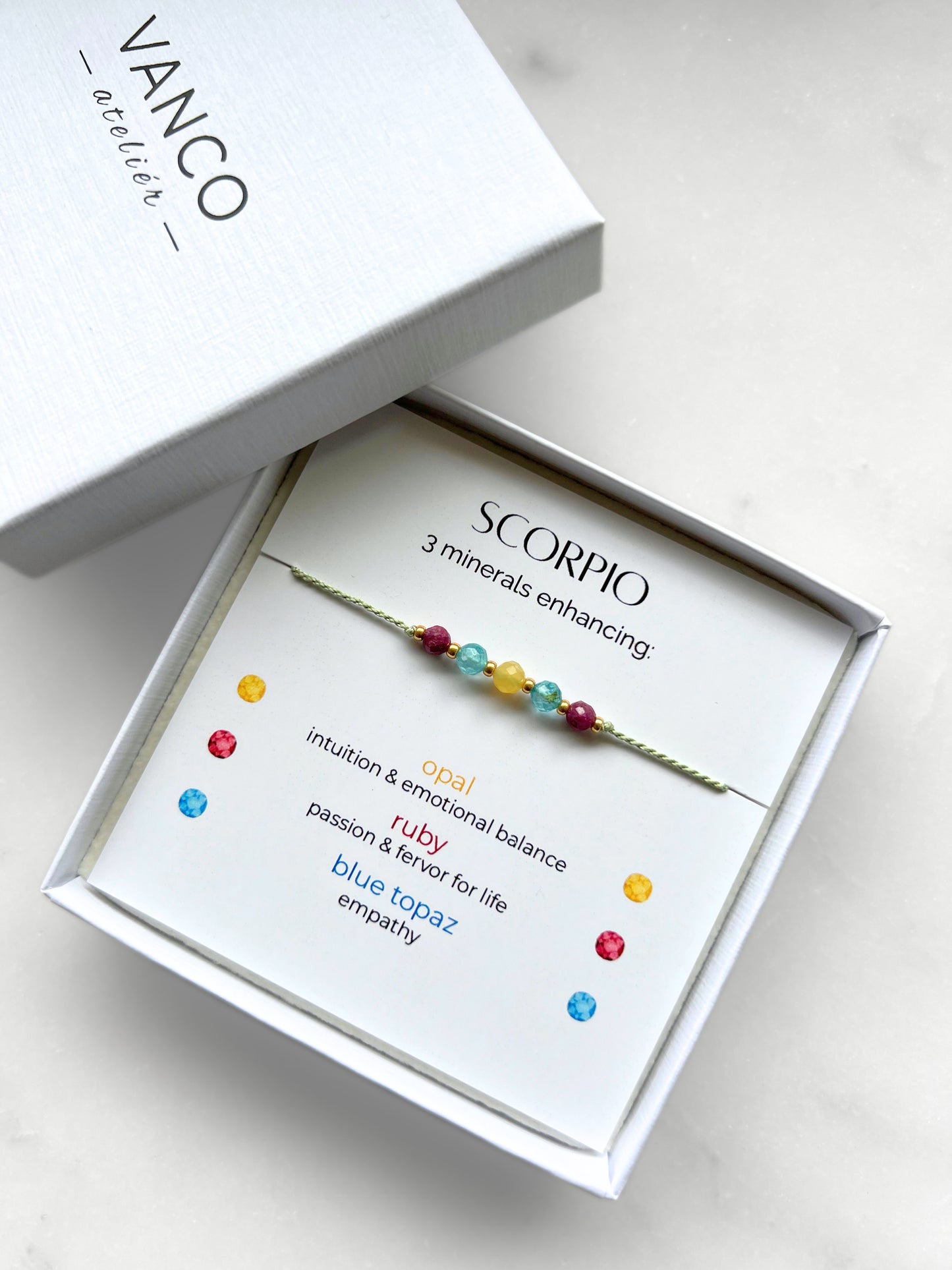 White gift box that contains a mineral bracelet for a Scorpio Zodiac sign. The bracelet is displayed on a card that describes the minerals and their meanings. There are five minerals on the bracelet: Two rubies, two blue topazes and one opal.