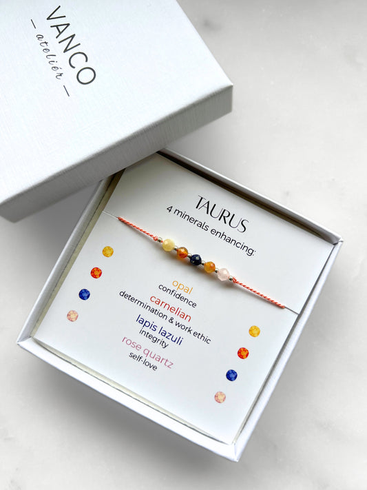 White gift box that contains a mineral bracelet for a Taurus Zodiac sign. The bracelet is displayed on a card that describes the minerals and their meanings. There are five minerals on the bracelet: opal, two carnelians, lapis lazuli and rose quartz