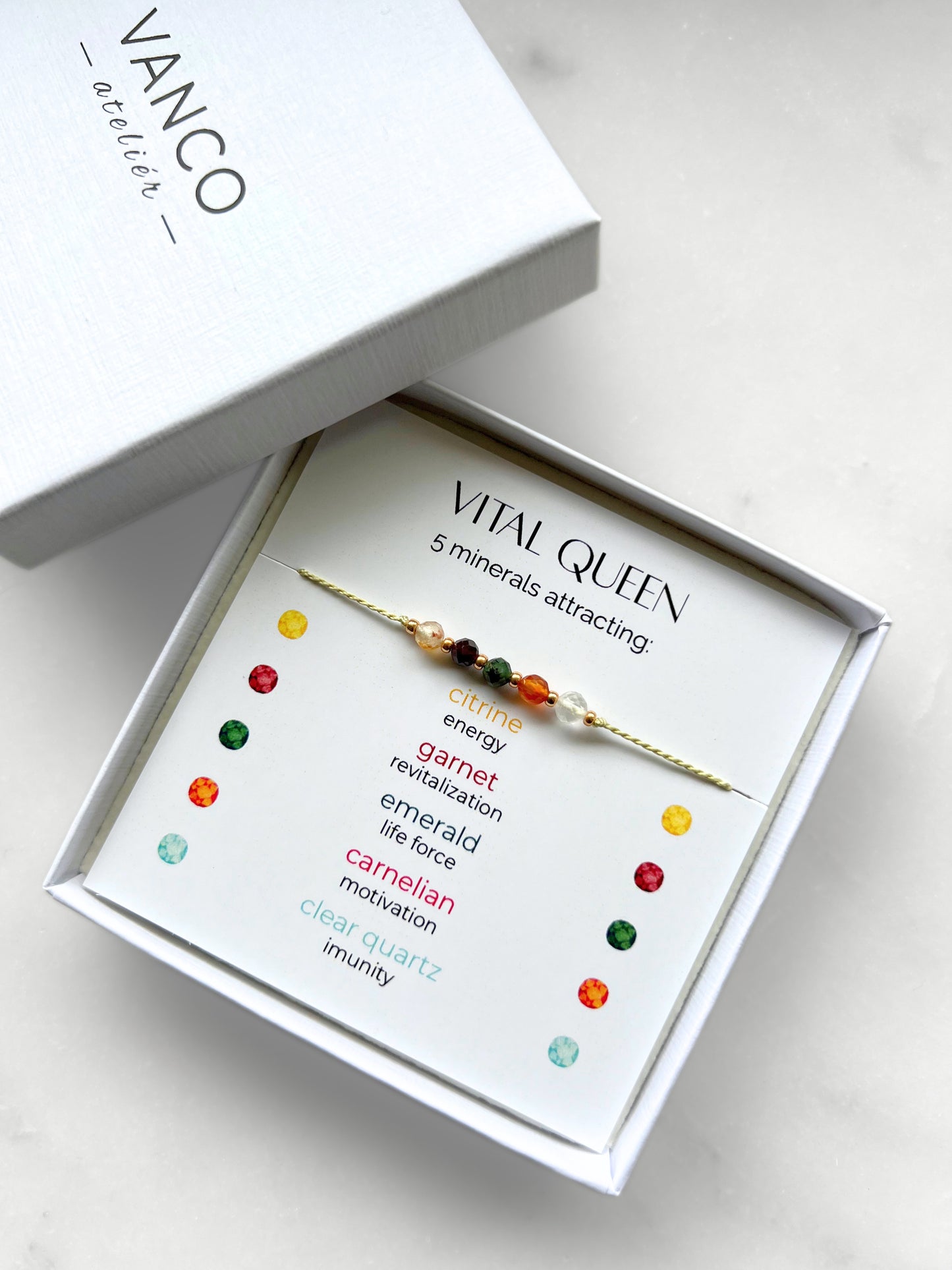 A white gift box that contains a bracelet made of green silk string with five mineral beads, put on a promotional card. The bracelet's theme is Vitality, the name of the bracelet is Vital Queen, which is written in the promotional card. The card also explains each stone and what each stone represents: Citrine for Energy, Garnet for Revitalization, Emerald for Life Force, Carnelian for Motivation, Clear Quartz for Imunity