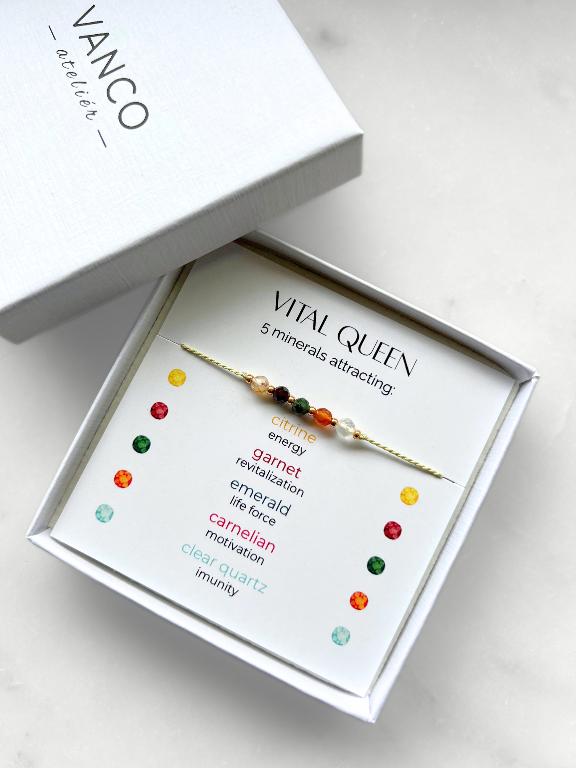 A white gift box that contains a bracelet made of green silk string with five mineral beads, put on a promotional card. The bracelet's theme is Vitality, the name of the bracelet is Vital Queen, which is written in the promotional card. The card also explains each stone and what each stone represents: Citrine for Energy, Garnet for Revitalization, Emerald for Life Force, Carnelian for Motivation, Clear Quartz for Imunity