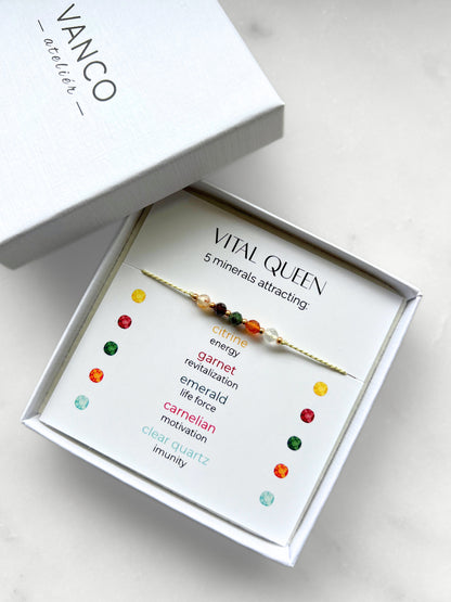 A white gift box that contains a bracelet made of green silk string with five mineral beads, put on a promotional card. The bracelet's theme is Vitality, the name of the bracelet is Vital Queen, which is written in the promotional card. The card also explains each stone and what each stone represents: Citrine for Energy, Garnet for Revitalization, Emerald for Life Force, Carnelian for Motivation, Clear Quartz for Imunity