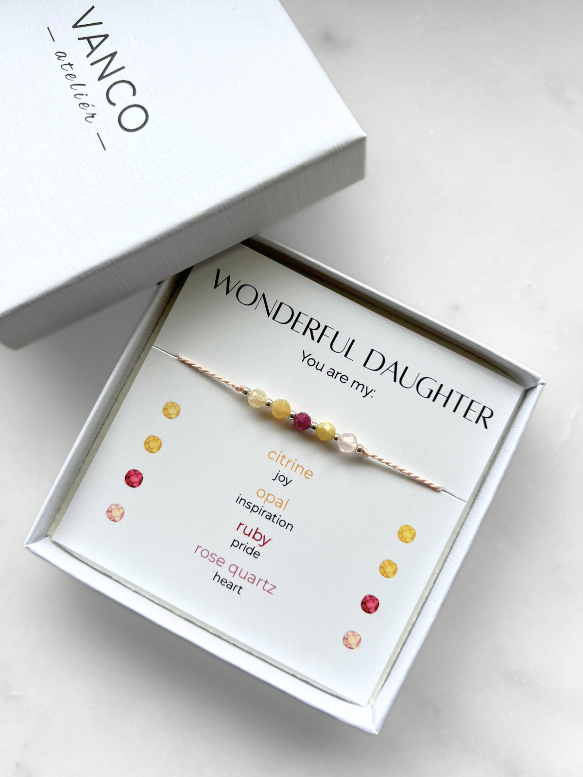 A white gift box that contains a bracelet made of pink silk string with five mineral beads, put on a promotional card. The name of the bracelet is "Wonderful daughter", which is written in the promotional card. The card also explains each stone and what message each stone sends to the recipient, saying "Your are my": Citrine for Joy, Opal for Inspiration, Ruby for Pride, and Rose Quartz for Heart.