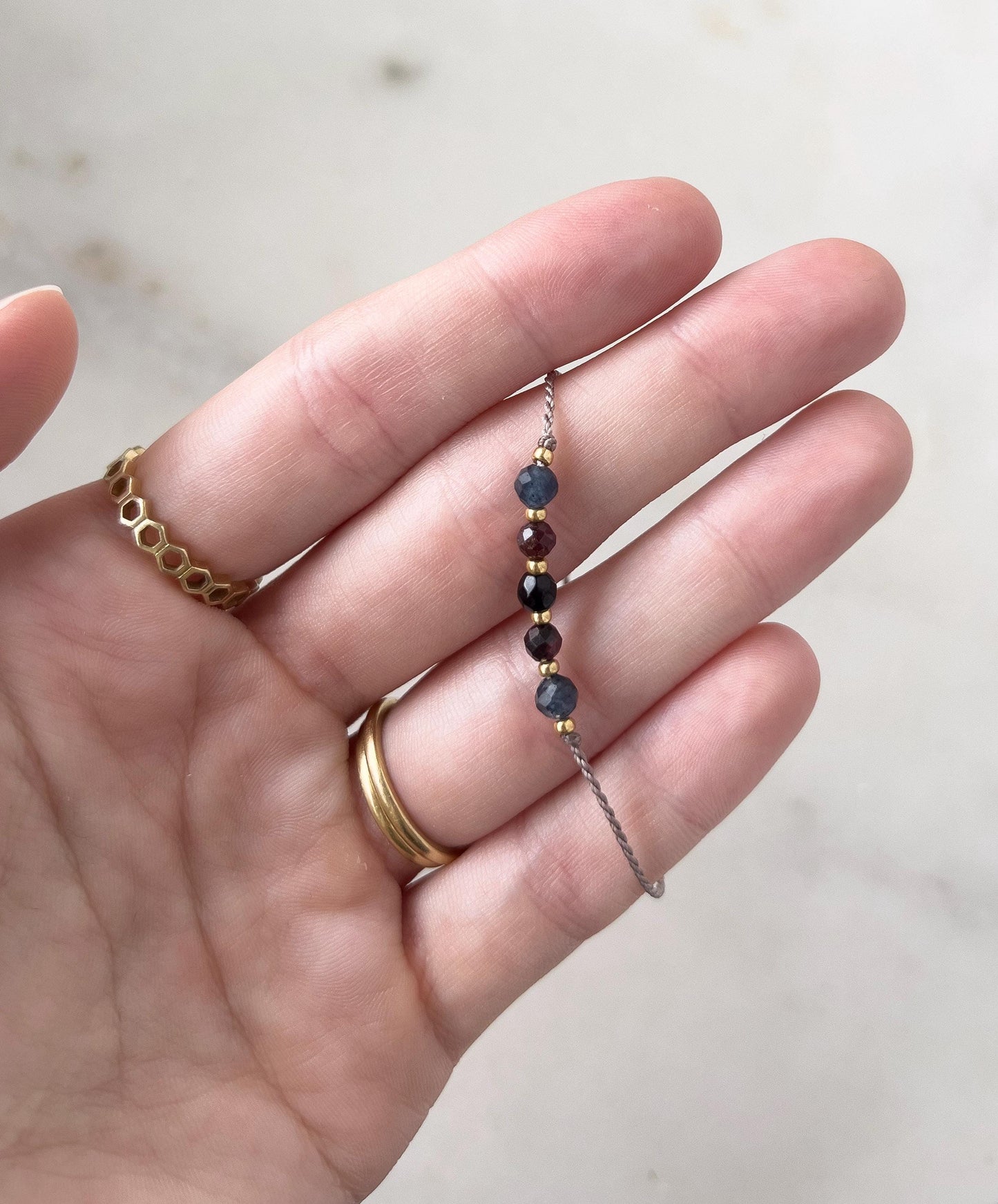 A delicate beaded bracelet with small, colourful gemstones. The bracelet features dark blue, burgundy, and gold-coloured beads. The bracelet is held in a hand, with gold rings on the fingers.