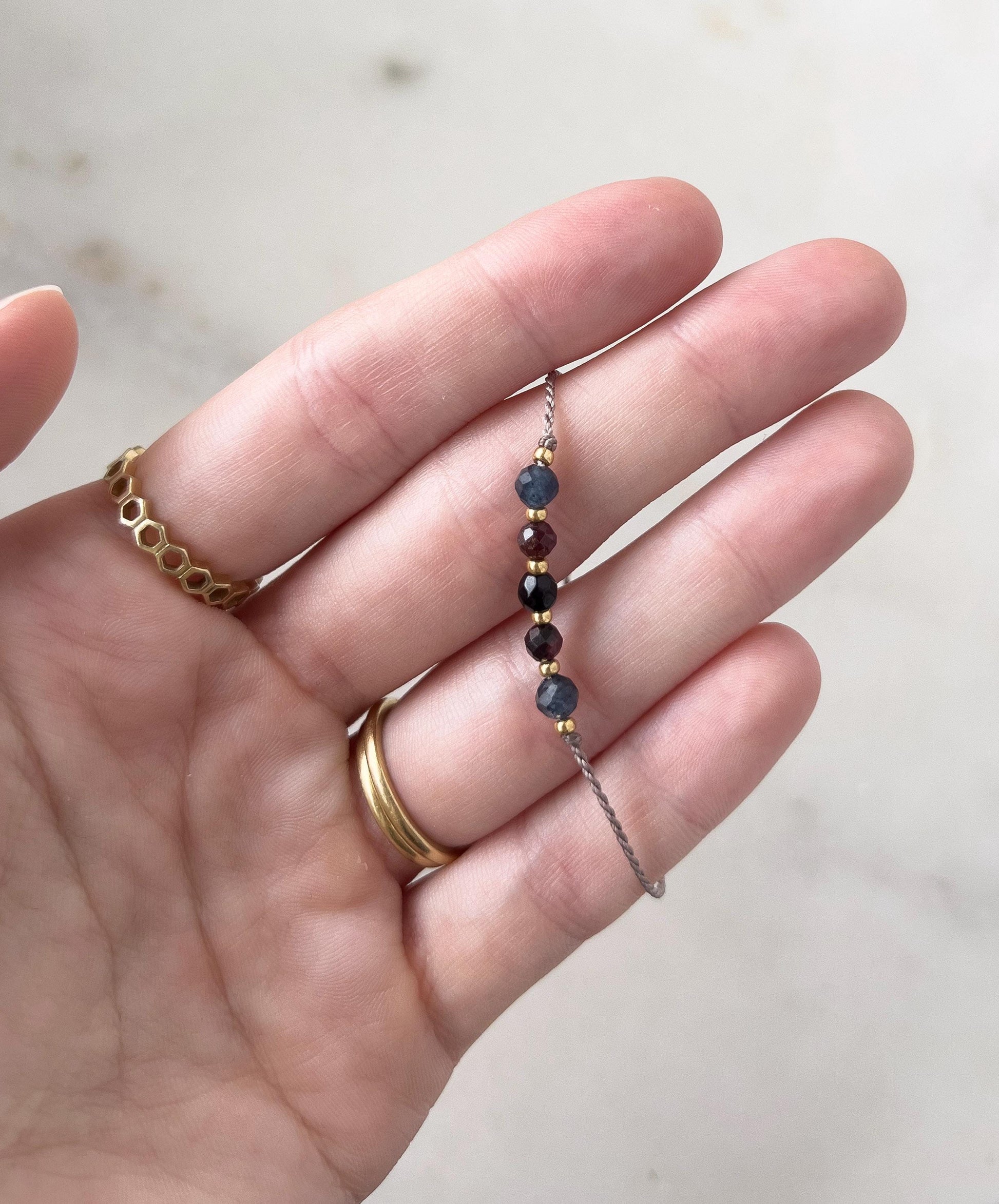 A delicate beaded bracelet with small, colourful gemstones. The bracelet features dark blue, burgundy, and gold-coloured beads. The bracelet is held in a hand, with gold rings on the fingers.