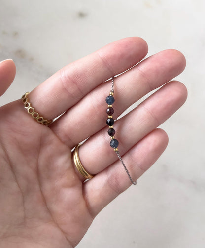 A delicate beaded bracelet with small, colourful gemstones. The bracelet features dark blue, burgundy, and gold-coloured beads. The bracelet is held in a hand, with gold rings on the fingers.