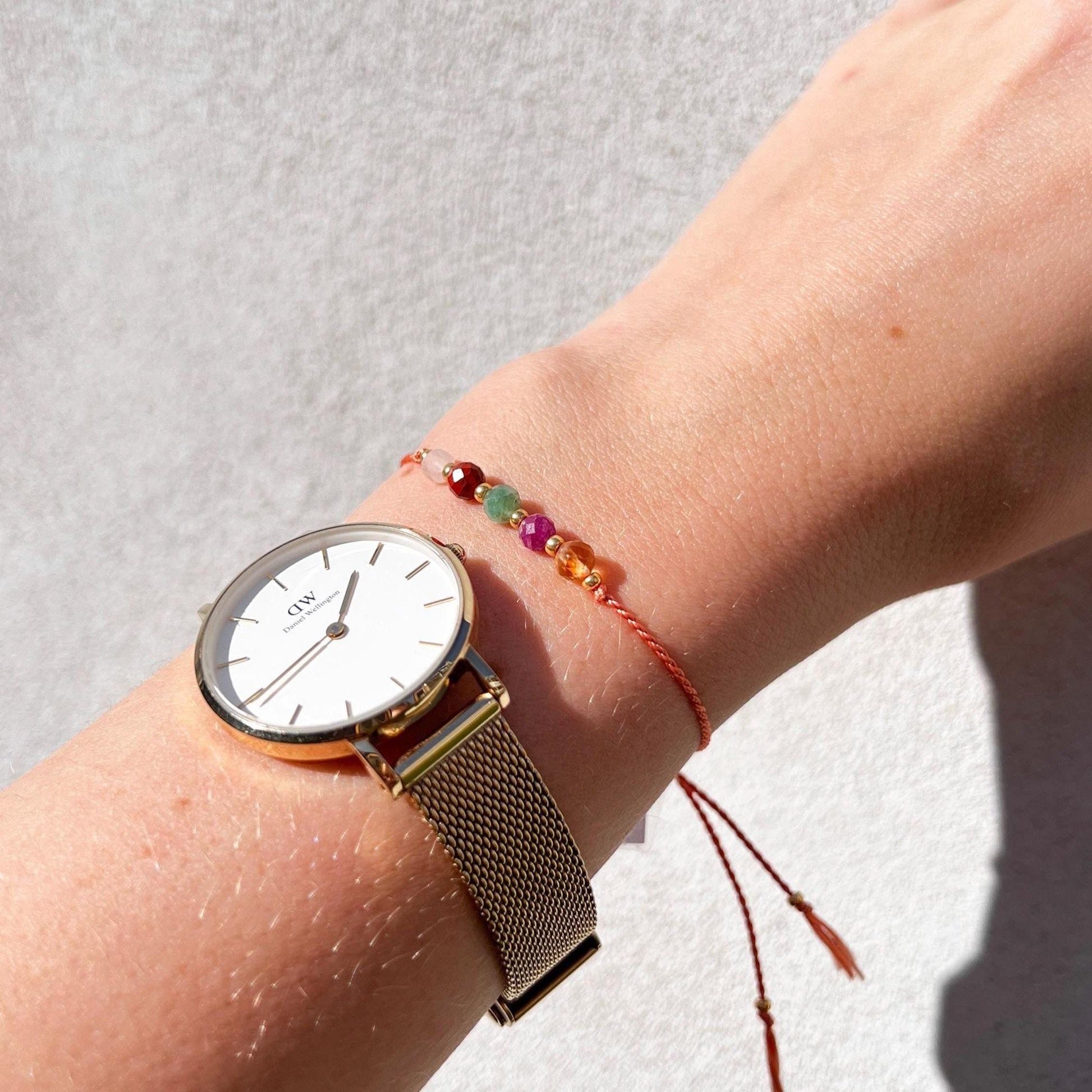 A female hand modeling a tangerine color silk cord bracelet strung with five mineral beads: white moonstone, red jasper, green emerald, pink ruby, yellow citrine. Each mineral is separated from another with a little gold bead, making the design more elegant. Model is also wearing a classic DW round watch with gold metal strap, showing how the bracelet can be a lovely addition to wear and layer with a watch.