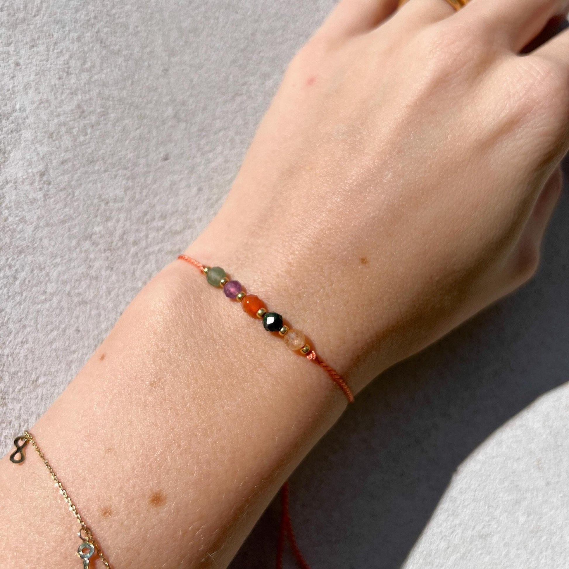 A female hand modeling a tangerine color silk cord bracelet strung with five mineral beads: yellow citrine, green jade, orange carnelian, purple amethyst, green aventurine. Each mineral is separated from another with a little gold bead, making the design more elegant. Model is also wearing a dainty gold bracelet together with the mineral bracelet, showing how the two can be elegantly layered together.
