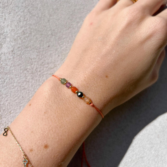 A female hand modeling a tangerine color silk cord bracelet strung with five mineral beads: yellow citrine, green jade, orange carnelian, purple amethyst, green aventurine. Each mineral is separated from another with a little gold bead, making the design more elegant. Model is also wearing a dainty gold bracelet together with the mineral bracelet, showing how the two can be elegantly layered together.