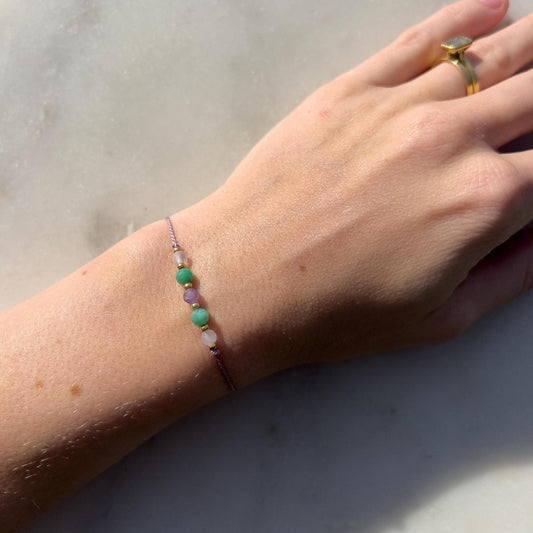 A female hand modeling a lilac silk cord bracelet strung with these mineral beads: white moonstone, blue amazonite, purple amethyst, another amazonite and another moonstone. Between the minerals, there are also little gold beads separating them and making the design more elegant.