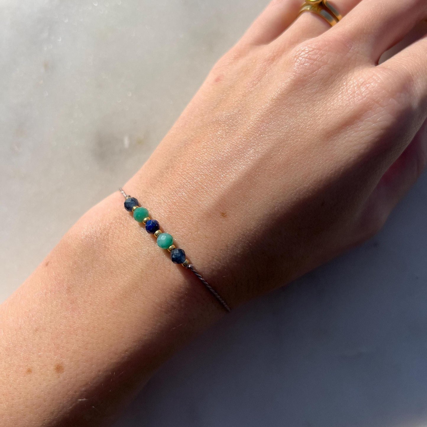 A female hand modeling a grey silk cord bracelet strung with these mineral beads: sapphire, amazonite, lapis lazuli, another amazonite and another sapphire. Between the minerals, there are also little gold beads separating them and making the design more elegant.