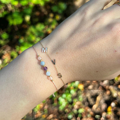 A female hand modeling a pink silk cord bracelet strung with 5 mineral beads: rose quartz, blue aquamarine, purple ametyst, another aquamarine and another rose quartz, with extra small gold beads between each mineral. Above this bracelet, model also wears gold initial bracelet to complete and layer the look.