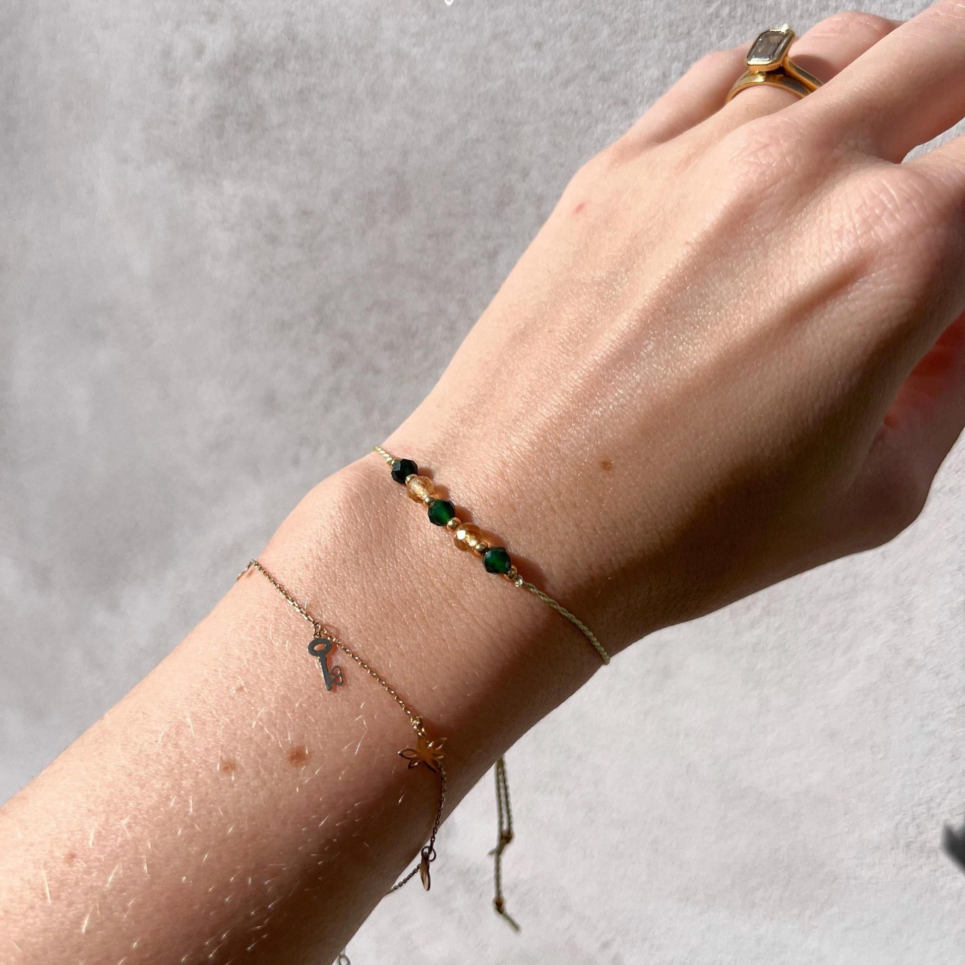 A female hand modeling a green silk cord bracelet strung with five mineral beads: alternating three green jades, two yellow citrines. Each mineral is separated from another with a little gold bead, making the design more elegant. Model is also wearing a dainty gold bracelet together with the mineral bracelet, showing how the two can be elegantly layered together.