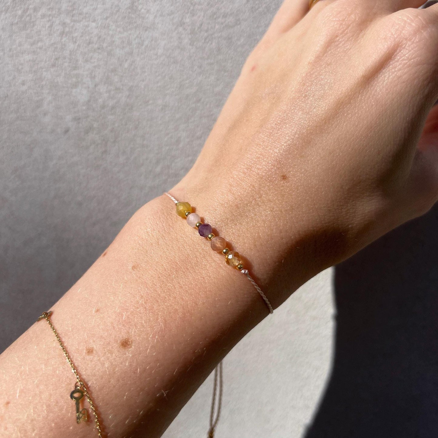 Dívčí ruka, která má na sobě tenký růžový hedvábný náramek s těmito minerály: žlutý citrín, sluneční kámen, fialový ametyst, růženín, žlutý opál. Mezi minerály jsou umístěny malé zlaté korálky, které dodávají náramku eleganci.