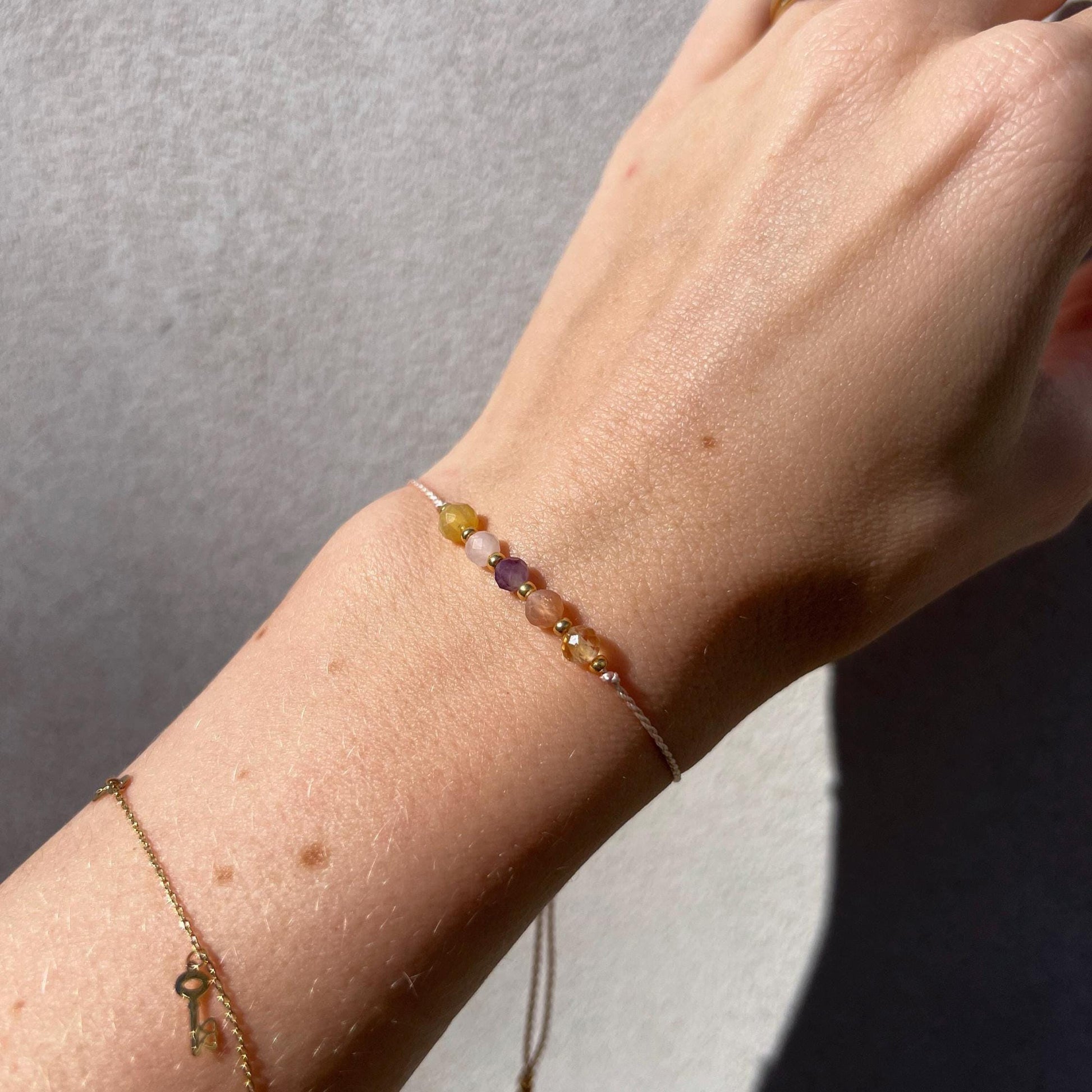 Dívčí ruka, která má na sobě tenký růžový hedvábný náramek s těmito minerály: žlutý citrín, sluneční kámen, fialový ametyst, růženín, žlutý opál. Mezi minerály jsou umístěny malé zlaté korálky, které dodávají náramku eleganci.