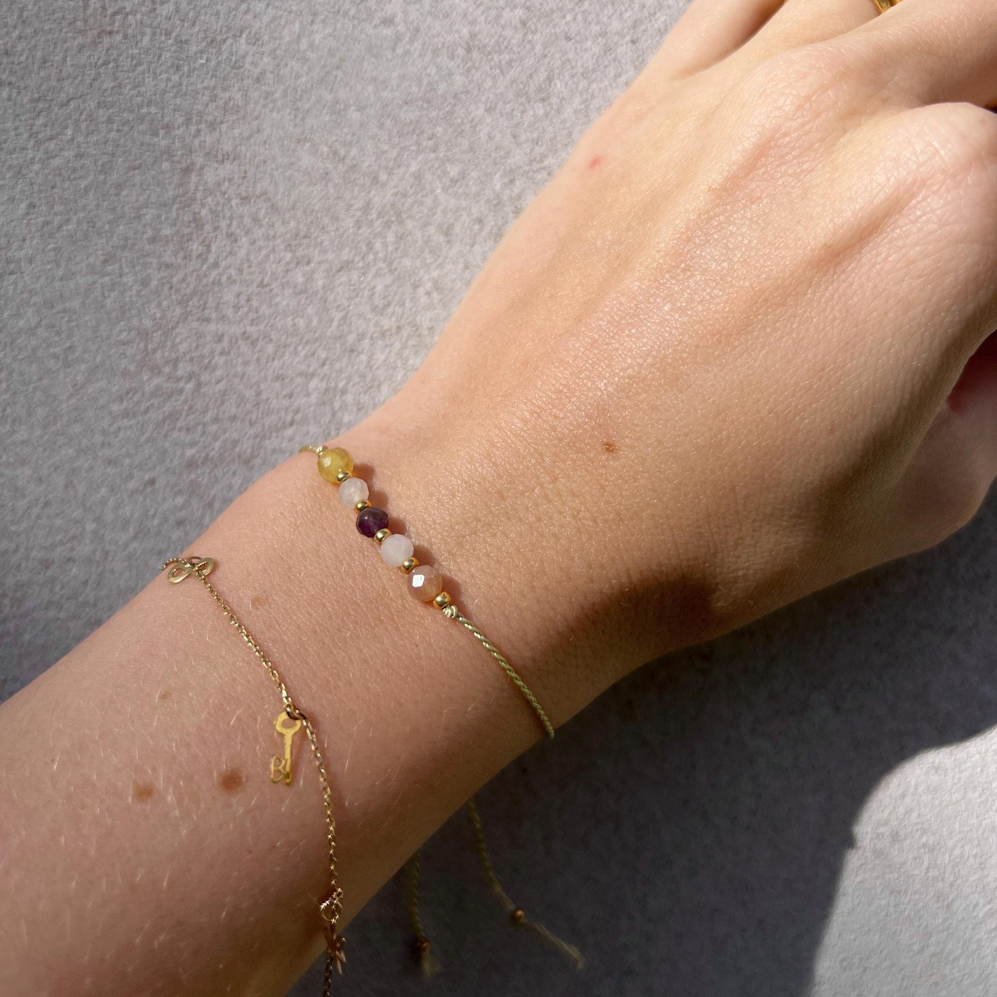 A female hand modeling a green silk cord bracelet strung with five mineral beads: purple amethyst, white moonstone, yellow opal, orange sunstone, rose quartz. Each mineral is separated from another with a little gold bead, making the design more elegant. Model is also wearing a dainty gold bracelet together with the mineral bracelet, showing how the two can be elegantly layered together.