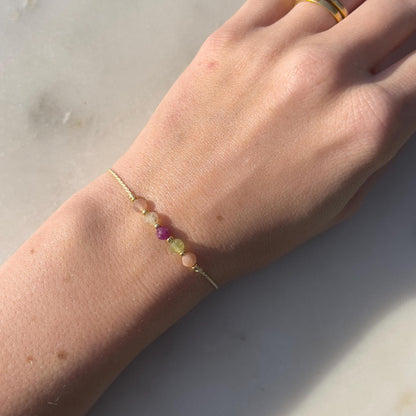 A female hand modeling a green silk cord bracelet strung with these mineral beads: orange sunstone, green peridot, red ruby, yellow citrine and another sunstone. Between the minerals, there are also little gold beads separating them and making the design more elegant.