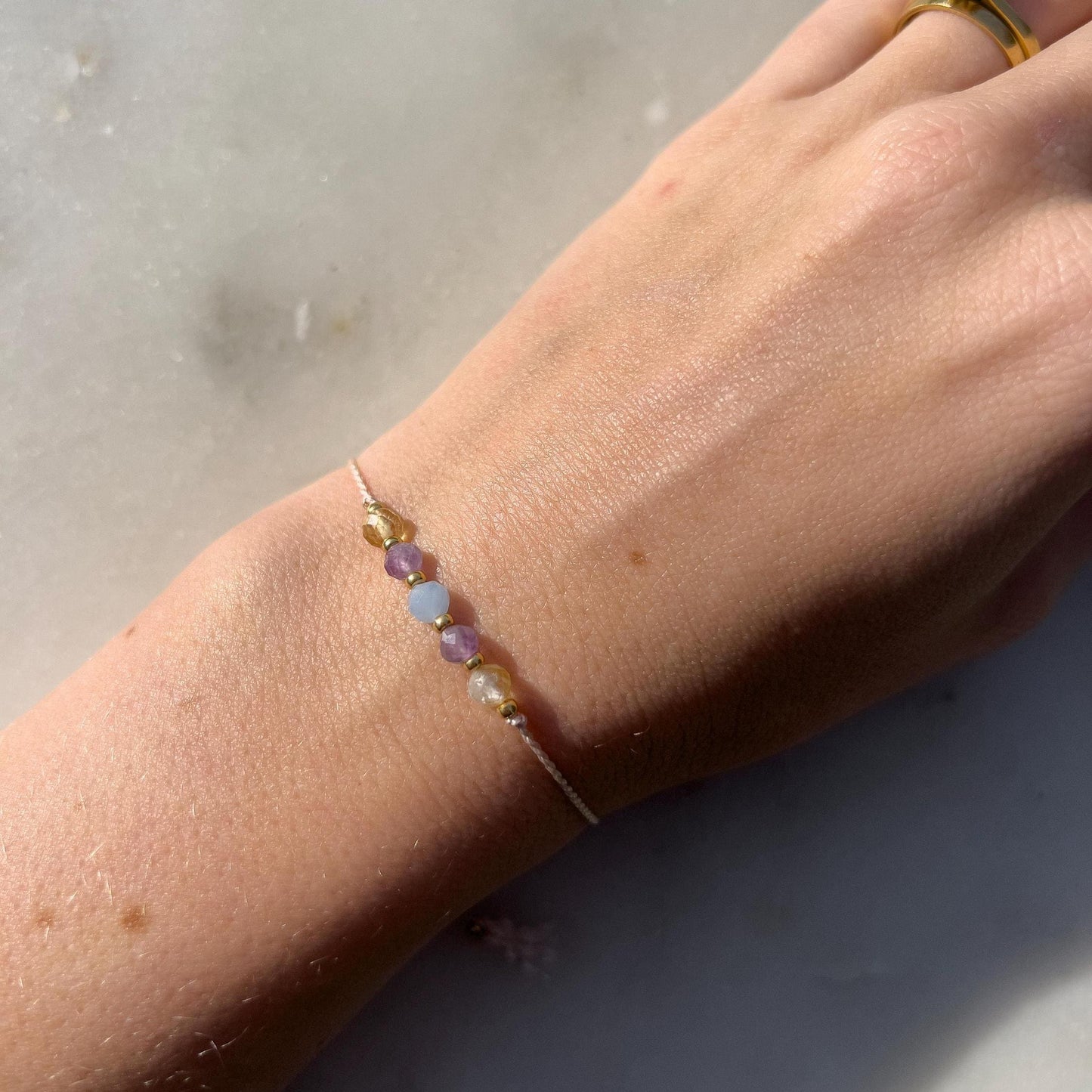 A female hand modeling a pink silk cord bracelet strung with these mineral beads: yellow citrine, purple amethyst, blue aquamarine, another amethyst and another citrine. Between the minerals, there are also little gold beads separating them and making the design more elegant.