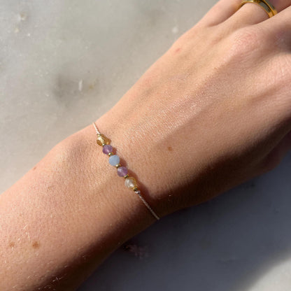 A female hand modeling a pink silk cord bracelet strung with these mineral beads: yellow citrine, purple amethyst, blue aquamarine, another amethyst and another citrine. Between the minerals, there are also little gold beads separating them and making the design more elegant.