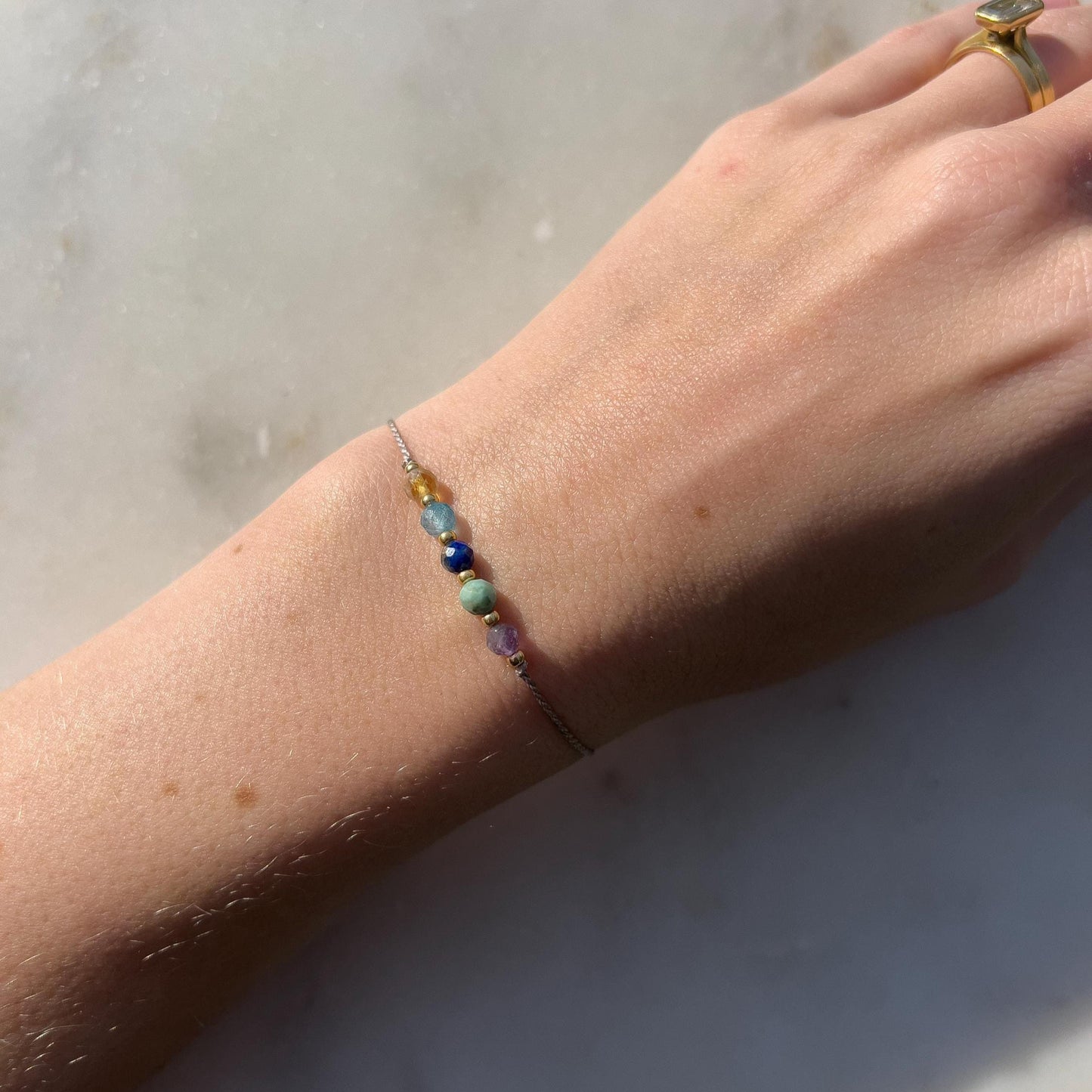 A hand modeling a grey silk cord bracelet strung with these mineral beads: purple amethyst, green turquoise, blue lapis lazuli, blue topaz and yellow citrine. Between the minerals, there are also little gold beads separating them and making the design more elegant.
