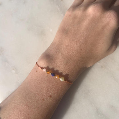 A female hand modeling a tangerine color silk cord bracelet strung with these mineral beads: yellow opal, orange carnelian, blue lapis lazuli, another carnelian and rose quartz. Between the minerals, there are also little gold beads separating them and making the design more elegant.