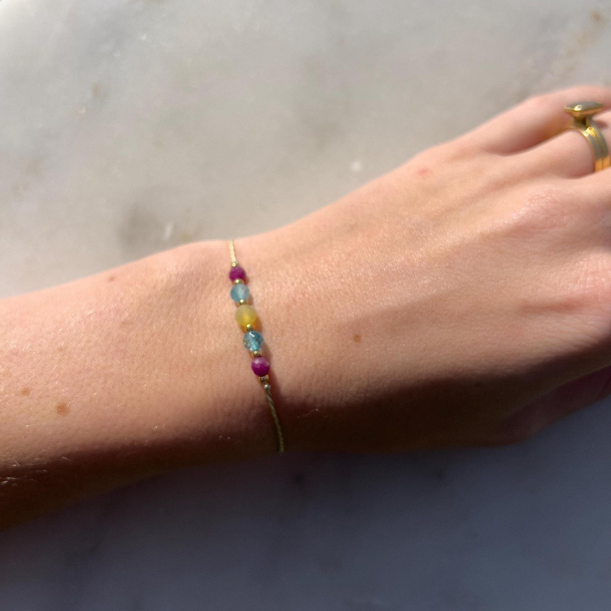 A hand modeling a green silk cord bracelet strung with these mineral beads: red ruby, blue topaz, yellow opal, another blue topaz and another ruby. Between each mineral is also a little gold bead separating the minerals and making the design more elegant.