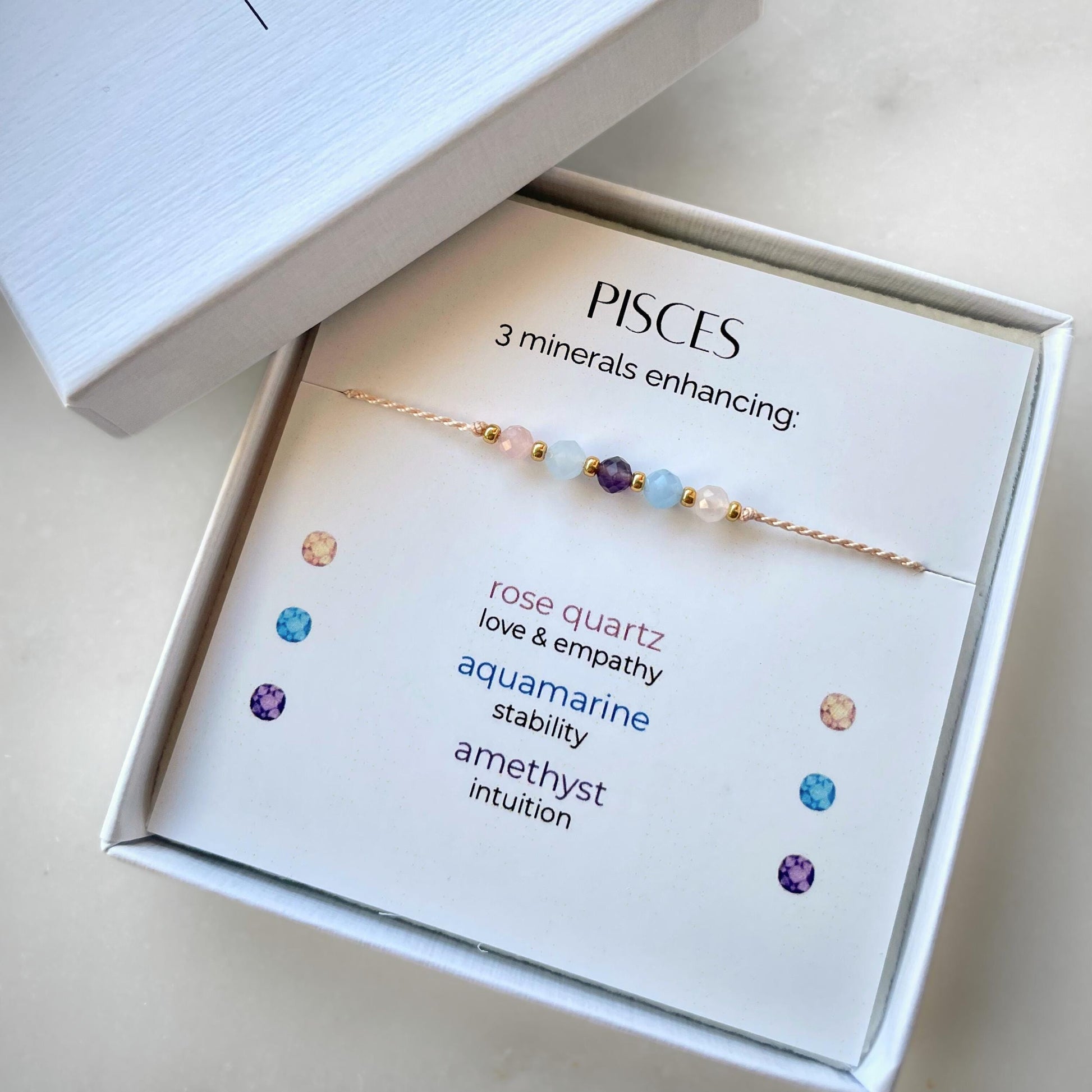 White gift box that contains a mineral bracelet for a Pisces Zodiac sign. The bracelet is displayed on a card that describes the minerals and their meanings. There are five minerals on the bracelet: two rose quartz, two aquamarines and amethyst