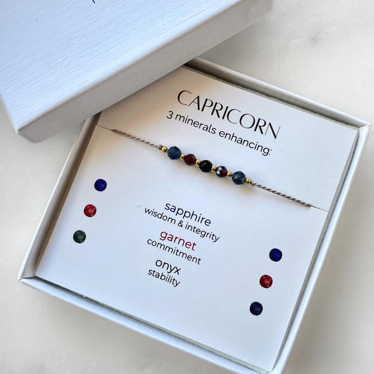 White gift box that contains a mineral bracelet for a Capricorn Zodiac sign. The bracelet is displayed on a card that describes the minerals and their meanings. There are five minerals on the bracelet: two sapphires, two garnets and onyx